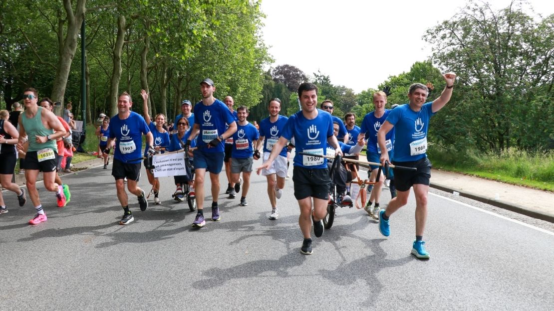 Een groep lopers in blauw shirt van Handicap International duwen twee personen in joëlettes voort