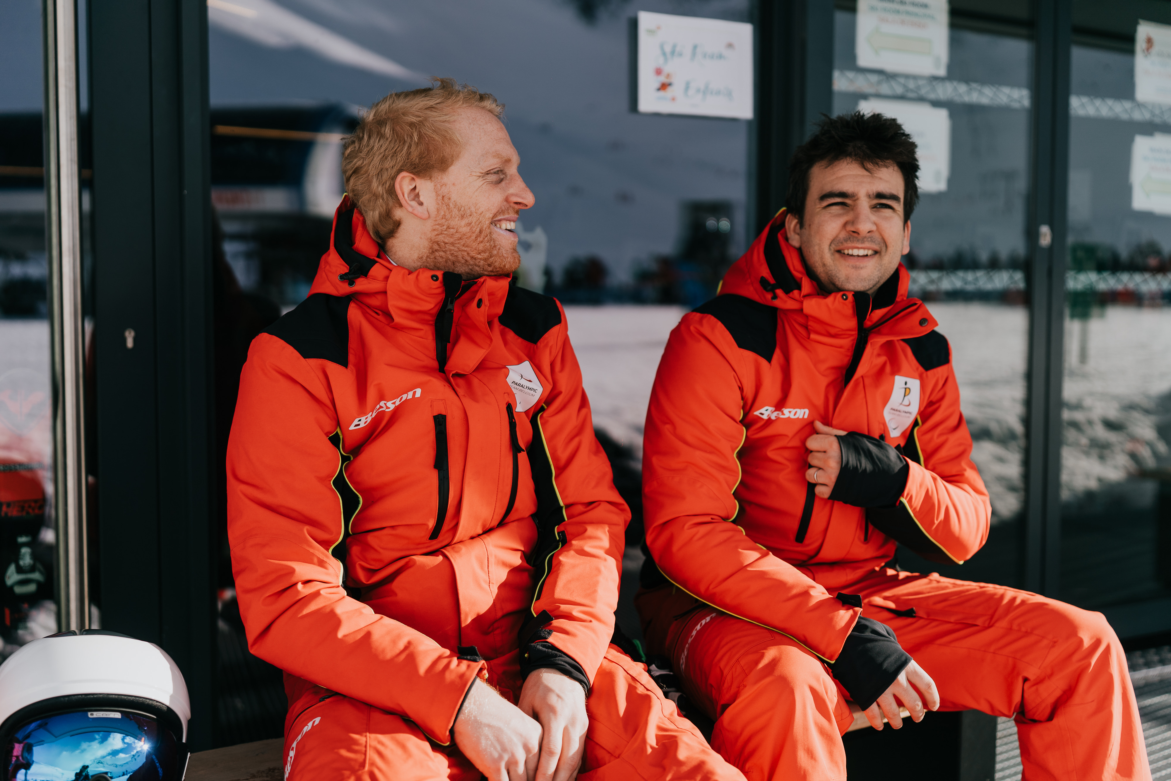 Maximilien Seeger et Jérémy Mestdagh, en tenue de ski, sont assis sur un banc et sourient. Ils sont adossés à une porte vitrée. Dans le reflet de la vitre, on aperçoit un paysage enneigé devant eux.