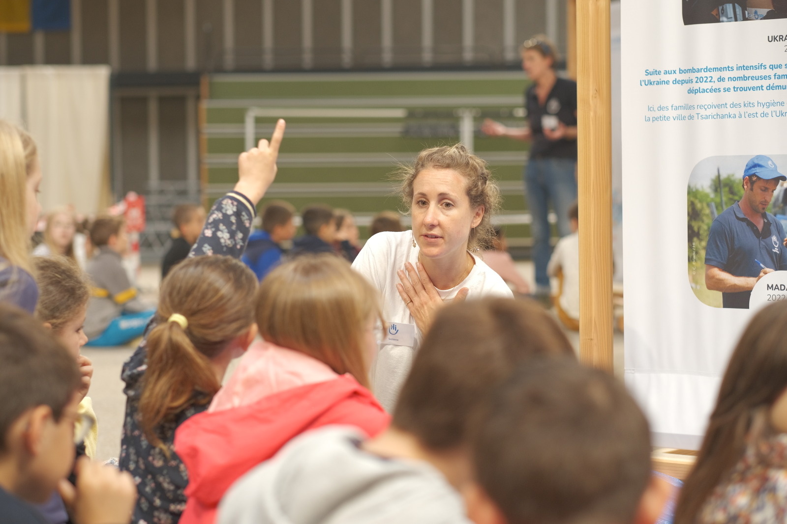 Een blonde vrouw staat voor een groep kinderen die met hun rug naar haar toe staan. Een klein meisje steekt haar hand op om een vraag te stellen.