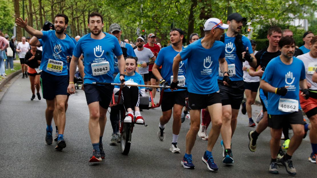 Un groupe de coureurs, revêtus de t-shirts bleus arborant le logo de Handicap International, se relayent pour pousser une joëlette.