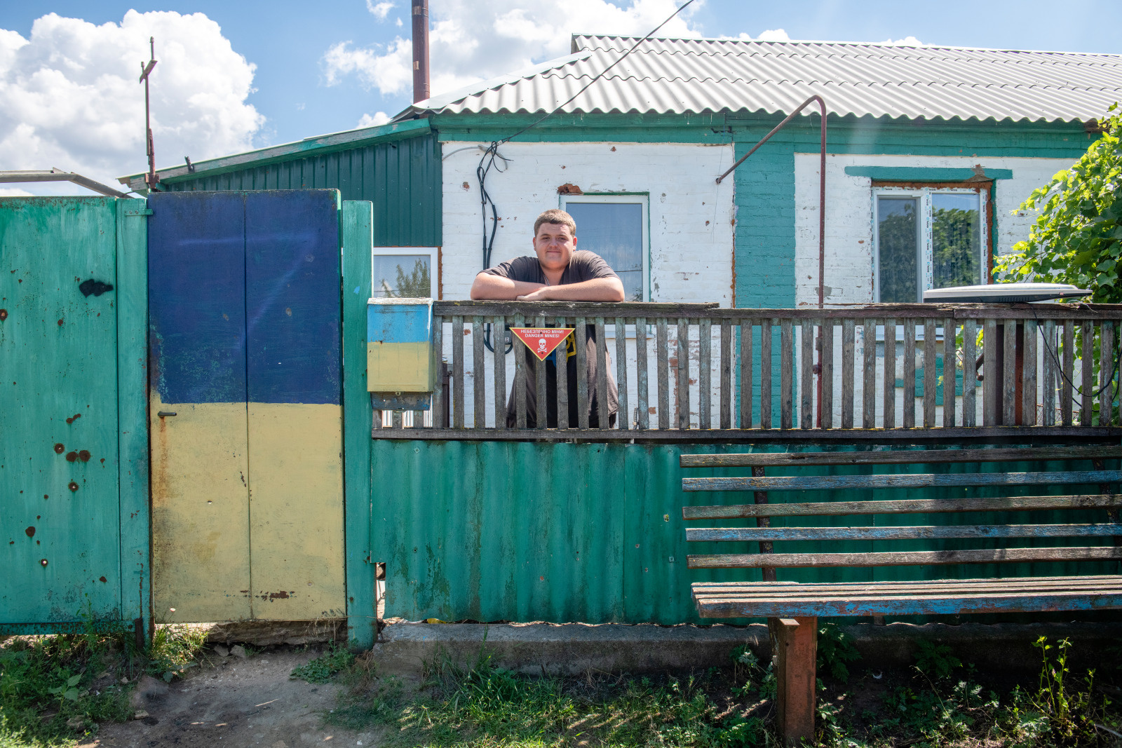 Un jeune homme se tient dans le jardin de devant de sa maison. Un petit panneau avertit de la présence de mines.