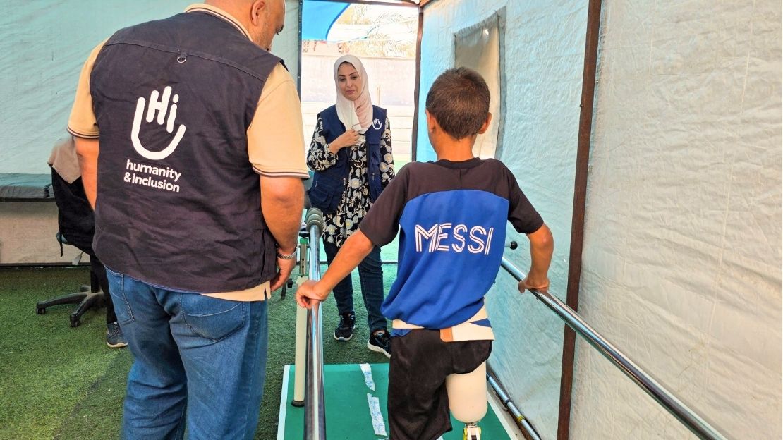 Een jongen met een beenprothese staat recht en houdt zich vast aan twee metalen baren. Twee personen met een jasje van Handicap International geven instructies.