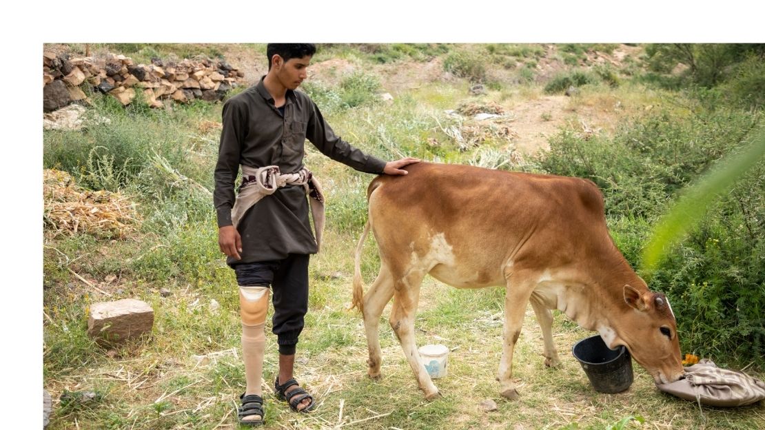 Een jongeman met een prothese staat naast een koe in een bergachtig landschap
