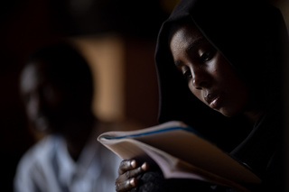 Une personne, vêtue d’un voile, lit attentivement un livre ou un cahier dans un environnement faiblement éclairé.