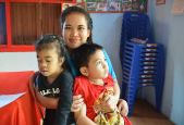 Une jeune femme prend dans ses bras deux enfants, une fille et un garçon. Latsaphone, enseignante formée par Handicap International qui accompagne des enfants comme Junior, 8 ans (à droite) dans une classe transitoire dans la province de Champassak, Laos.  