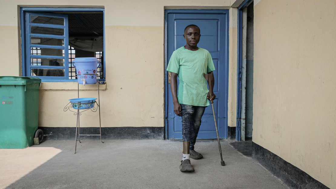 Un homme se tient debout, appuyé sur une béquille. La jambe droite de son pantalon est relevée pour découvrir une prothèse. Derrière lui, un mur avec une fenêtre et deux portes.