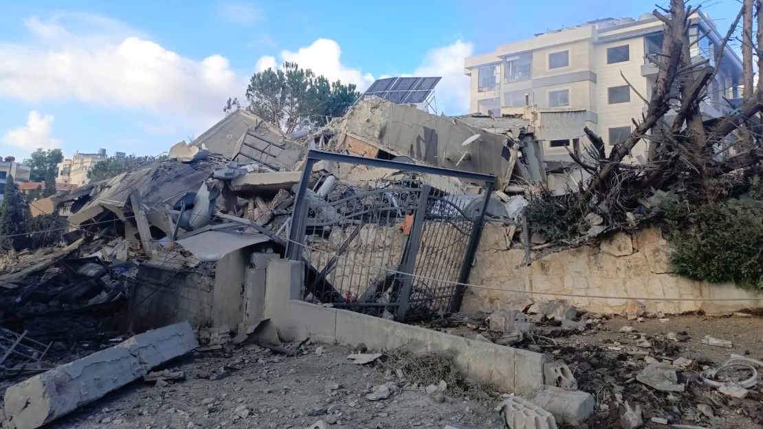 De foto toont een ingestort gebouw, met gebroken betonnen blokken, een vervormd metalen hek en beschadigde bomen op de voorgrond. Op de achtergrond zijn andere gebouwen nog intact, onder een bewolkte hemel.