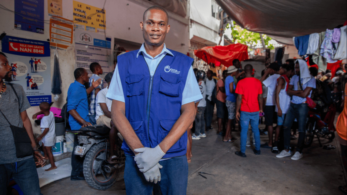 Portrait d'un homme souriant portant des gants de travail et une jaquette Handicap International. Derrière lui, on devine une salle où discutent de nombreuses personnes et où sèche du linge.