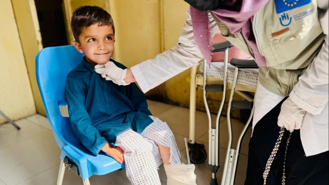 Un petit garçon souriant est assis sur une chaise et regarde une femme penchée sur lui qui porte une jaquette HI / Union européenne.