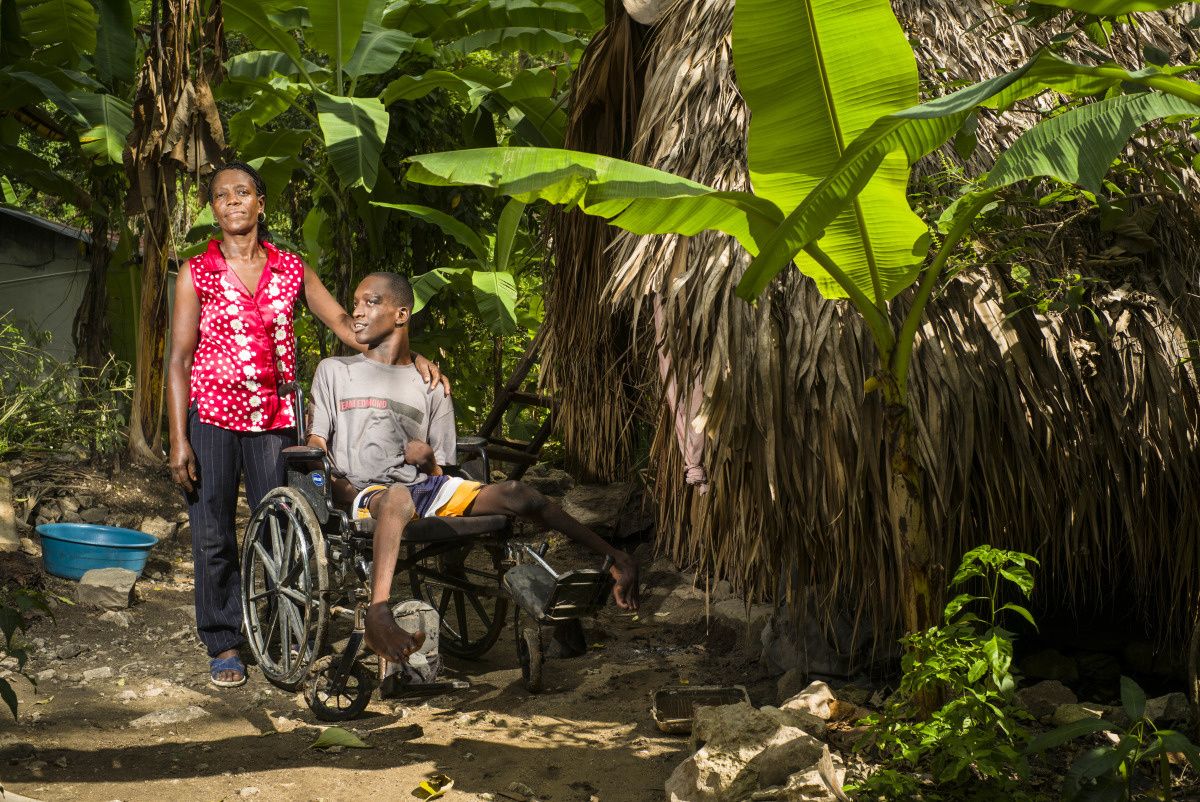 Jerilia, une main posée sur l'épaule de son fils, assis dans sa chaise roulante. Ils se trouvent sur un chemin de terre, entourés de palmiers.