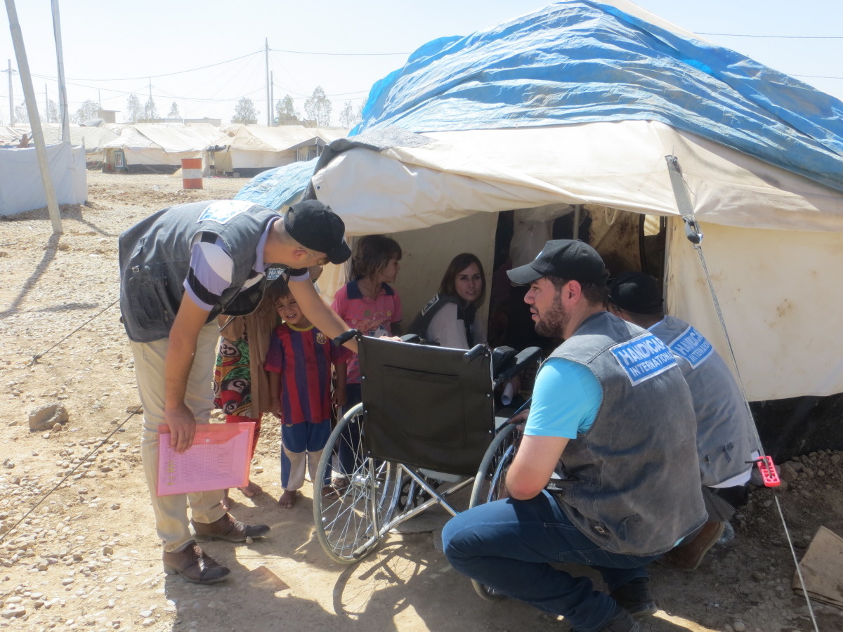 Bij een tent in een ontheemdenkamp geven medewerkers van Handicap International een rolstoel af aan een familie. 