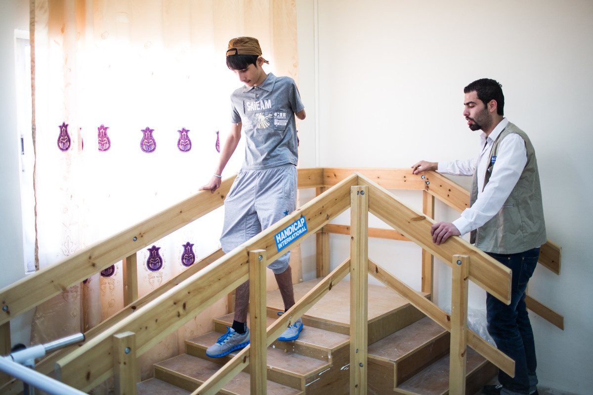 Un adolescent sans bras gauche, un fixateur externe sur une jambe descend d'un petit escalier d'exercice en bois dans une salle de kinésithérapie. Un kinésithérapeute de Handicap International regarde le garçon. 