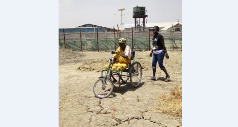 Mary sur le tricycle donné par Handicap International. Une dame marche derrière elle. Le sol est desséché.
