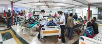 Une salle de l'hôpital remplie de lits occupés, deux personnes de l'équipe de Handicap International se penchent sur un patient alité