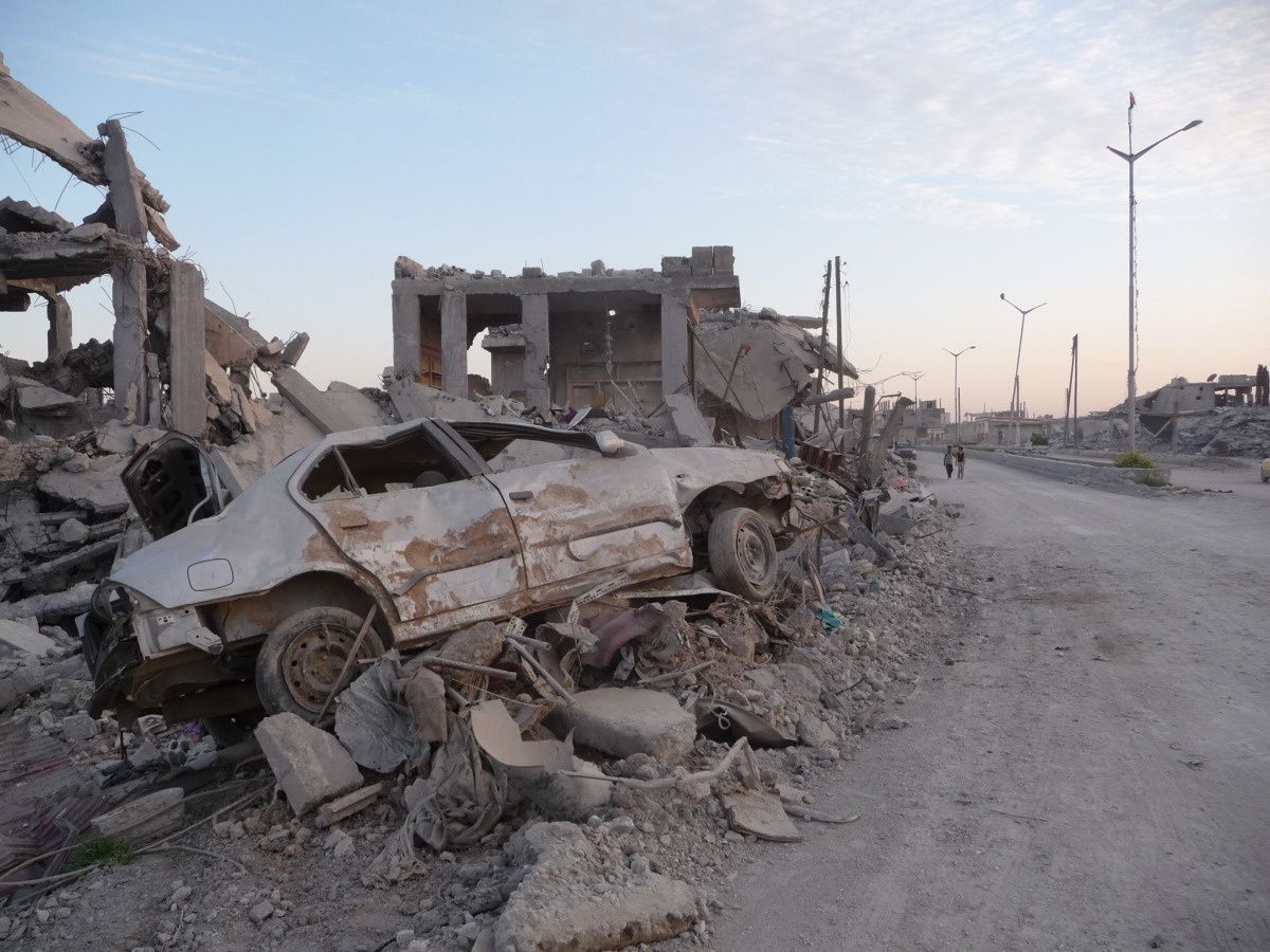 Une rue de Kobané. Tous les batiments alentours sont détruits, une carcasse de voiture couchée sur le flanc, sur des décombres, se trouve à l'avant-plan.